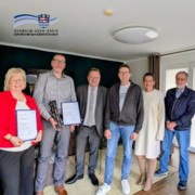 Sechs Erwachsene stehen in einem Wohnraum nebeneinander und blicken in die Kamera. Zwei Personen halten gerahmte Urkunden in den Händen, eine weitere Person hält eine dekorative Auszeichnung. Die Gruppe ist gepflegt gekleidet, teils in Business-, teils in Alltagskleidung. Oben links ist das Logo des Schwalm-Eder-Kreises eingeblendet.