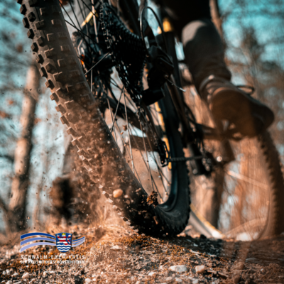 Zu sehen ist eine Nahaufnahme eines Mountainbike-Hinterrads auf einem Waldweg. Das Profil des Reifens ist deutlich erkennbar, während beim Fahren Erde und kleine Steine aufgewirbelt werden. Im Hintergrund sind unscharf Bäume zu sehen. Unten links ist das Logo des Schwalm-Eder-Kreises eingeblendet.