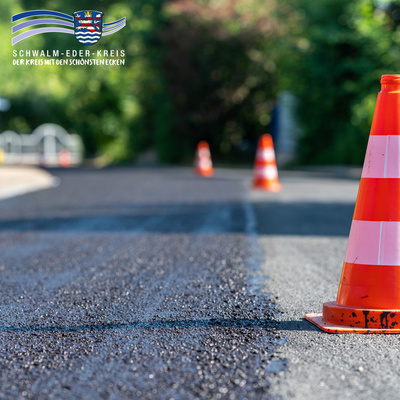 Nahaufnahme einer frisch asphaltierten Straße mit mehreren orange-weißen Warnkegeln, die den Arbeitsbereich absichern. Die Sonne spiegelt sich auf dem neuen Asphalt, im Hintergrund sind Bäume und Büsche zu sehen. Oben links ist das Logo des Schwalm-Eder-Kreises mit dem Schriftzug „Der Kreis mit den schönsten Ecken“ eingeblendet.