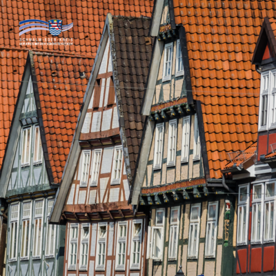 Das Bild zeigt eine Reihe historischer Fachwerkhäuser mit steilen, orangefarbenen Ziegeldächern. Die Häuser stehen eng beieinander und unterscheiden sich in Farbe und Muster der Holzbalken. Die weißen Fenster heben sich kontrastreich von den dunklen Balken ab. Oben links im Bild ist das Logo des Schwalm-Eder-Kreises mit dem Schriftzug „Der Kreis mit den schönsten Ecken“ zu sehen.