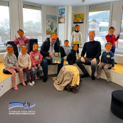 In einem hellen Raum mit großen Fenstern sitzen mehrere Kinder und zwei Erwachsene auf einer gepolsterten Sitzbank. Die Kinder tragen Tiger-Masken vor dem Gesicht. Einer der Männer hält ein Kinderbuch in die Kamera. Im Hintergrund hängen bunte Poster an den Wänden. Unten links ist das Logo des Schwalm-Eder-Kreises zu sehen.
