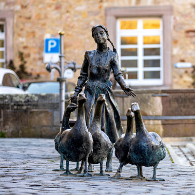 Bronzeskulptur „Gänseliesel“ in Melsungen: Eine junge Frau mit geflochtenem Zopf, gekleidet in ein schlichtes Kleid, steht auf einem gepflasterten Platz und wird von fünf Gänsen umringt. Die Figur hält eine Hand leicht nach vorne ausgestreckt, als würde sie die Tiere führen. Im Hintergrund sind ein sandsteinfarbenes Gebäude mit weißen Fenstern, ein Straßenschild und ein geparktes Auto zu sehen.