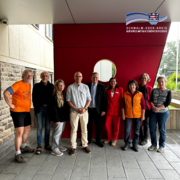Neun Personen stehen gemeinsam vor einer roten Wand mit dem Logo des Schwalm-Eder-Kreises. Die Gruppe posiert für ein Foto vor einem Gebäude. Die Personen tragen unterschiedliche Kleidung, darunter Freizeitkleidung, eine orangefarbene Weste und einen roten Hosenanzug. In der Mitte stehen zwei Personen in formeller Kleidung. Die Aufnahme entstand im überdachten Eingangsbereich des Gebäudes.