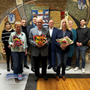 Auf dem Bild steht eine Gruppe von Frauen und Männern in einem Innenraum der Kreisverwaltung Schwalm-Eder. Mehrere Personen halten Blumensträuße in den Händen. Sie haben sich zu Ehrungen und Verabschiedungen zusammengefunden. Im Hintergrund ist eine große Wand mit einem Mosaik zu sehen, das Wappen und Ortsnamen des Schwalm-Eder-Kreises zeigt. Die Gruppe steht ruhig nebeneinander und blickt in Richtung Kamera.