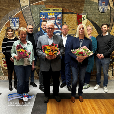 Auf dem Bild steht eine Gruppe von Frauen und Männern in einem Innenraum der Kreisverwaltung Schwalm-Eder. Mehrere Personen halten Blumensträuße in den Händen. Sie haben sich zu Ehrungen und Verabschiedungen zusammengefunden. Im Hintergrund ist eine große Wand mit einem Mosaik zu sehen, das Wappen und Ortsnamen des Schwalm-Eder-Kreises zeigt. Die Gruppe steht ruhig nebeneinander und blickt in Richtung Kamera.