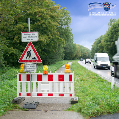 Eine rot-weiße Absperrung mit gelben Warnleuchten versperrt einen Rad- und Fußweg entlang einer Landstraße. Auf den Schildern steht: „Fußgänger bitte andere Straßenseite benutzen“ und „Radfahrer bitte andere Straßenseite benutzen“. Rechts fahren mehrere Autos auf der nassen Fahrbahn, links ist dichter, grüner Wald zu sehen. Oben rechts befindet sich das Logo des Schwalm-Eder-Kreises mit dem Schriftzug „Der Kreis mit den schönsten Ecken“.