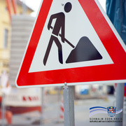 Nahaufnahme eines rot-weißen Baustellenschilds mit dem Symbol eines Arbeiters, der mit einer Schaufel Erde bewegt. Im Hintergrund sind unscharf Absperrungen und Baustellenmaterialien zu erkennen. Unten rechts ist das Logo des Schwalm-Eder-Kreises mit dem Schriftzug „Der Kreis mit den schönsten Ecken“ eingeblendet.