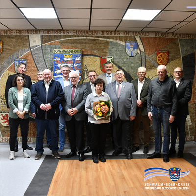 Auf einem Gruppenfoto in der Kreisverwaltung Homberg stehen elf Personen vor einer großen Wappen- und Landkreiskarte an der Wand. Im Vordergrund hält eine Frau einen bunten Blumenstrauß, daneben und hinter ihr stehen mehrere Männer in Anzügen und in Freizeitkleidung. Unten rechts ist das Logo des Schwalm-Eder-Kreises zu sehen; das Bild entstand im Rahmen der Verleihung der Ehrenbriefe des Landes Hessen.