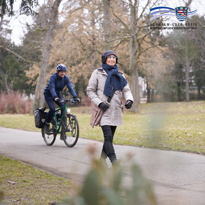 Eine Frau geht lächelnd auf einem Gehweg durch einen Park. Sie trägt eine helle Winterjacke, einen blauen Schal, Mütze, Handschuhe und eine Umhängetasche. Hinter ihr fährt ein Mann auf einem grünen E-Bike. Er trägt dunkle Kleidung und einen Fahrradhelm. Es ist Winter – die Bäume sind kahl, der Rasen grün-braun. Im Bild oben rechts ist das Logo des Schwalm-Eder-Kreises zu sehen.