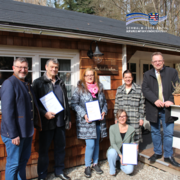 Sechs Erwachsene stehen und knien vor einem Holzgebäude im Außenbereich und blicken in die Kamera. Drei von ihnen halten gerahmte Urkunden in den Händen. Im Hintergrund sind Fenster, eine Außenlampe sowie ein Schild am Gebäude zu sehen. Oben rechts im Bild ist das Logo des Schwalm-Eder-Kreises mit dem Schriftzug „Der Kreis mit den schönsten Ecken“ eingeblendet.
