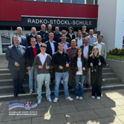 Gruppenfoto vor dem Gebäude der Radko-Stöckl-Schule: Mehrere Erwachsene sowie vier Jugendliche stehen gemeinsam auf einer Treppe und blicken in die Kamera. Die Jugendlichen in der ersten Reihe halten kleine technische Bauteile mit Kabeln in den Händen. Hinter ihnen stehen weitere Personen in Business- und Alltagskleidung. Über dem Eingang ist der Schriftzug „Radko-Stöckl-Schule“ zu sehen. Unten links im Bild befindet sich das Logo des Schwalm-Eder-Kreises.