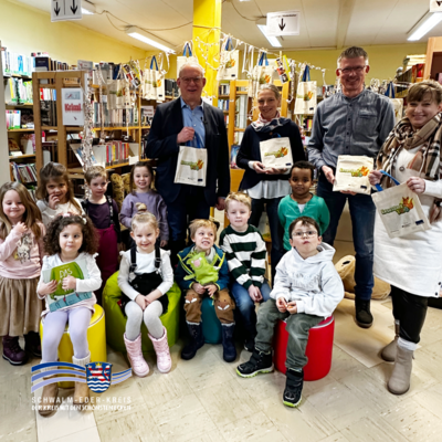 In einer Bibliothek stehen mehrere Erwachsene und Kinder zwischen Bücherregalen. Einige Erwachsene halten Stofftaschen mit der Aufschrift „Lesestart 1–2–3“ in die Kamera. Vor ihnen sitzen Kinder auf bunten Hockern, teilweise mit Büchern in der Hand. Im Hintergrund sind Regale mit Büchern und aufgehängte Taschen zu sehen. Unten links ist das Logo des Schwalm-Eder-Kreises abgebildet.