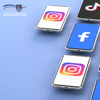 Mehrere moderne Smartphones liegen auf einer hellblauen Oberfläche. Auf ihren Bildschirmen sind bekannte Social-Media-Logos zu sehen: Instagram (mehrfach), Facebook und TikTok. Links oben im Bild ist das Logo des Schwalm-Eder-Kreises mit dem Schriftzug „Der Kreis mit den schönsten Ecken“ eingeblendet. Die Darstellung symbolisiert die Nutzung sozialer Medien.