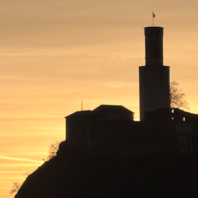 Silhouette einer Burg auf einem Hügel vor einem orange-goldenen Sonnenuntergang. Der dunkle Umriss zeigt einen runden Turm und mehrere Gebäude, rechts stehen einzelne Bäume, der Himmel ist von warmen Abendfarben durchzogen.