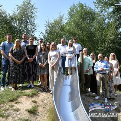 Eine Gruppe von 16 Personen steht im Freien vor Bäumen bei sonnigem Wetter. Alle schauen lächelnd in die Kamera. Sie posieren neben einer silberfarbenen Kinderrutsche. Rechts unten im Bild ist das Logo des Schwalm-Eder-Kreises mit der Aufschrift: „Schwalm-Eder-Kreis – Der Kreis mit den schönsten Ecken“. Auf dem Bild zu sehen: Von links: Jan-Lukas Girth, Lena Dörrbecker, Lars Beckmann, Emily Macht, Lucas Dickhaut, Judith Kurz, Mirlinta Cermjani, Julia Kemmerling, Winfried Becker (Landrat), Lara Eckhardt, Tobias Heipel (Personalleiter), Ange-lika Roth (Personalrat), Yuma Klitsch (JAV), René Petzold, Sina Sämmler (Ausbildungsleiterin), Gerlinde Eckhardt (Frauen- und Gleichstellungsbeauftragte)
