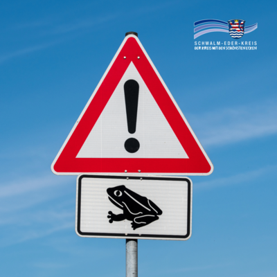 Zu sehen ist ein Verkehrsschild vor blauem Himmel. Oben befindet sich ein dreieckiges Warnschild mit rotem Rand und einem schwarzen Ausrufezeichen. Darunter ist ein Zusatzschild mit der schwarzen Silhouette eines Frosches angebracht. Rechts oben im Bild steht das Logo des Schwalm-Eder-Kreises mit Schriftzug.