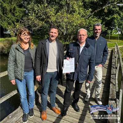 Vier Personen stehen auf einer hölzernen Brücke über einem kleinen Fluss und schauen in die Kamera. Von links: Babette Tanner (Fachbereich Bauen und Umwelt in der Kreisverwaltung), Bürgermeister Achim Siebert, Erster Kreisbeigeordneter Jürgen Kaufmann (mit einem Dokument in der Hand) und Gerhard Reuter (Fachbereichsleiter Bauen und Umwelt). Im Hintergrund sind Bäume und eine Wiese zu sehen. Unten rechts im Bild befindet sich das Logo des Schwalm-Eder-Kreises mit Wappen und der Aufschrift: „Schwalm-Eder-Kreis – Der Kreis mit den schönsten Ecken“.