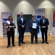 Vier Personen stehen in einem Raum mit Holzfußboden nebeneinander und blicken in die Kamera. In der Mitte hält eine Frau ein offizielles Dokument mit dem Hessen-Wappen in der Hand. Links und rechts stehen je ein Mann im Anzug, links außen eine Frau in heller Jacke. Im Hintergrund sind zwei Roll-Up-Banner mit Logos des Landes Hessen und des Europäischen Sozialfonds Plus sowie eine Hessen-Flagge zu sehen. Es handelt sich um eine feierliche Übergabe oder Auszeichnung. Auf dem Bild von links: Staatssekretärin Manuela Strube, Lars Werner (Fachbereichsleiter Sozialverwaltung Schwalm-Eder), Sabine Feisel (Projekt Ready2Care) und Landrat Winfried Becker.