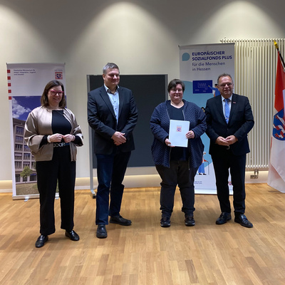 Vier Personen stehen in einem Raum mit Holzfußboden nebeneinander und blicken in die Kamera. In der Mitte hält eine Frau ein offizielles Dokument mit dem Hessen-Wappen in der Hand. Links und rechts stehen je ein Mann im Anzug, links außen eine Frau in heller Jacke. Im Hintergrund sind zwei Roll-Up-Banner mit Logos des Landes Hessen und des Europäischen Sozialfonds Plus sowie eine Hessen-Flagge zu sehen. Es handelt sich um eine feierliche Übergabe oder Auszeichnung. Auf dem Bild von links: Staatssekretärin Manuela Strube, Lars Werner (Fachbereichsleiter Sozialverwaltung Schwalm-Eder), Sabine Feisel (Projekt Ready2Care) und Landrat Winfried Becker.