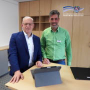 Zwei Männer stehen in einem Büro hinter einem Schreibtisch und blicken in die Kamera. Einer trägt einen blauen Anzug mit weißem Hemd, der andere ein grünes Hemd mit Namensschild. Auf dem Tisch steht ein Tablet mit Tastaturhülle, im Hintergrund sind helle Holzschränke zu sehen. Oben rechts ist das Logo des Schwalm-Eder-Kreises eingeblendet.