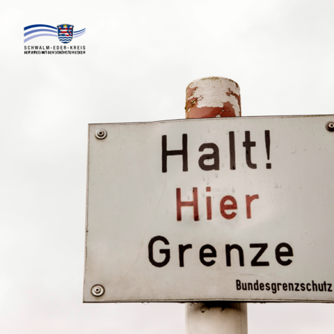 Auf dem Bild ist ein altes Metallschild an einem Pfosten zu sehen. Darauf steht gut lesbar „Halt! Hier Grenze“. Das Schild ist leicht verwittert und vor hellem Himmel fotografiert. Oben links befindet sich das Logo des Schwalm-Eder-Kreises.