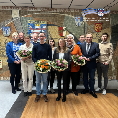 Mehrere Erwachsene stehen in einem Innenraum vor einer großen Wandgestaltung mit dem Schriftzug „Schwalm-Eder-Kreis“ und verschiedenen Wappen. Einige Personen in der vorderen Reihe halten Blumensträuße in den Händen. Die Gruppe blickt in die Kamera. Oben rechts ist das Logo des Schwalm-Eder-Kreises eingeblendet.