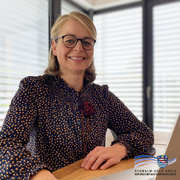Frau mit blonden Haaren und Brille sitzt lächelnd an einem Tisch. Sie trägt eine dunkelblaue Bluse mit gelblichen Punkten und einer roten Stoffblume als Schmuck. Im Hintergrund sind große Fenster mit geschlossenen Jalousien zu sehen.