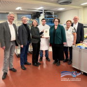Gruppenfoto in einer Wäscherei: Sieben Personen stehen zwischen Maschinen und Wäschewagen. In der Mitte wird eine Urkunde überreicht. Unten rechts ist das Signet des Schwalm-Eder-Kreises eingeblendet.