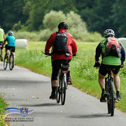 Mehrere Radfahrerinnen und Radfahrer fahren auf einem asphaltierten Radweg durch eine grüne Landschaft. Die Personen tragen Helme, Rucksäcke und sportliche Kleidung. Im Hintergrund sind Wiesen, Bäume und ein Waldrand zu sehen. Unten links befindet sich das Logo des Schwalm-Eder-Kreises mit dem Schriftzug „Der Kreis mit den schönsten Ecken“.