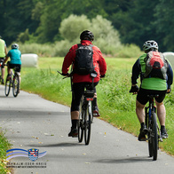 Mehrere Radfahrerinnen und Radfahrer fahren auf einem asphaltierten Radweg durch eine grüne Landschaft. Die Personen tragen Helme, Rucksäcke und sportliche Kleidung. Im Hintergrund sind Wiesen, Bäume und ein Waldrand zu sehen. Unten links befindet sich das Logo des Schwalm-Eder-Kreises mit dem Schriftzug „Der Kreis mit den schönsten Ecken“.