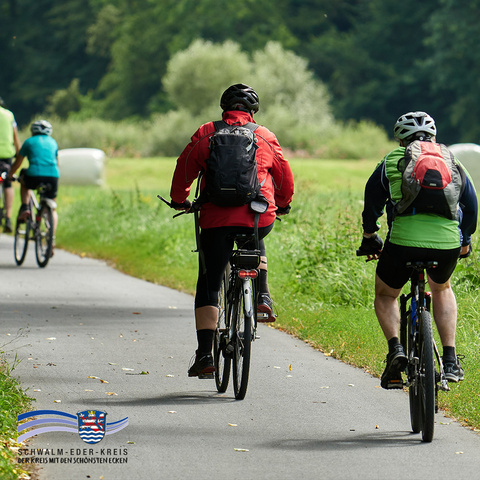 Mehrere Radfahrerinnen und Radfahrer fahren auf einem asphaltierten Radweg durch eine grüne Landschaft. Die Personen tragen Helme, Rucksäcke und sportliche Kleidung. Im Hintergrund sind Wiesen, Bäume und ein Waldrand zu sehen. Unten links befindet sich das Logo des Schwalm-Eder-Kreises mit dem Schriftzug „Der Kreis mit den schönsten Ecken“.