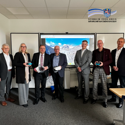 In einem Besprechungsraum stehen sieben Personen nebeneinander vor einer großen digitalen Tafel, auf der eine Landschaft mit Bergen zu sehen ist. Die Personen tragen formelle Kleidung und halten teilweise Virtual-Reality-Brillen und technische Geräte in den Händen. Der Raum wirkt modern und hell, mit Büromobiliar und Präsentationstechnik im Hintergrund. In der oberen rechten Ecke ist das Logo des Schwalm-Eder-Kreises zu sehen.