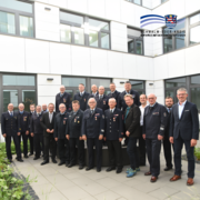 Gruppenfoto im Innenhof eines modernen Verwaltungsgebäudes: Mehr als 20 Personen stehen in mehreren Reihen und blicken in die Kamera. Ein Großteil der Anwesenden trägt Feuerwehruniformen mit Auszeichnungen, einige Personen sind in ziviler Kleidung gekleidet. Im Hintergrund sind die Fassade des Gebäudes mit großen Fenstern sowie ein bepflanzter Bereich zu sehen. Das Bild entstand im Rahmen einer Ehrungsveranstaltung für langjähriges Engagement im Feuerwehrwesen. Oben rechts befindet sich das Logo des Schwalm-Eder-Kreises.