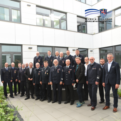 Gruppenfoto im Innenhof eines modernen Verwaltungsgebäudes: Mehr als 20 Personen stehen in mehreren Reihen und blicken in die Kamera. Ein Großteil der Anwesenden trägt Feuerwehruniformen mit Auszeichnungen, einige Personen sind in ziviler Kleidung gekleidet. Im Hintergrund sind die Fassade des Gebäudes mit großen Fenstern sowie ein bepflanzter Bereich zu sehen. Das Bild entstand im Rahmen einer Ehrungsveranstaltung für langjähriges Engagement im Feuerwehrwesen. Oben rechts befindet sich das Logo des Schwalm-Eder-Kreises.