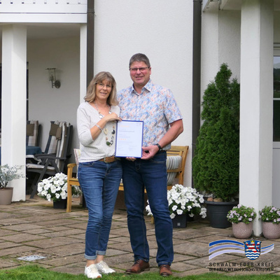 Claudia und Matthias Knoch stehen vor ihrem Haus im Garten und halten gemeinsam eine Urkunde in den Händen. Beide lächeln in die Kamera. Im Hintergrund sind Sitzmöbel, Blumenkübel und die Hausfassade zu sehen.