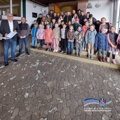 Gruppenfoto vor einem Gebäude: Eine Schulklasse mit vielen Kindern steht auf Treppenstufen, links daneben zwei Erwachsene, einer hält eine Urkunde. Unten rechts ist das Logo des Schwalm-Eder-Kreises eingeblendet.