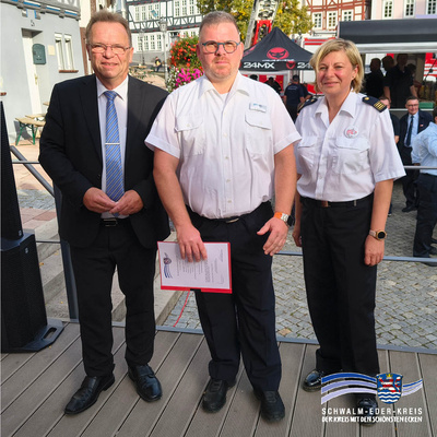 Landrat Winfried Becker, Björn Nöchel und Kreisbrandinspektorin Tanja Dittmar stehen nebeneinander und lächeln in die Kamera. Björn Nöchel hält eine Urkunde in der Hand. Im Hintergrund sind Fachwerkhäuser, ein rotes Zelt und weitere Menschen zu sehen. Unten rechts im Bild ist das Logo des Schwalm-Eder-Kreises mit dem Schriftzug „Der Kreis mit den schönsten Ecken“ abgebildet.