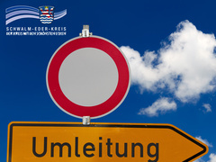 Zwei Verkehrsschilder vor blauem Himmel mit weißen Wolken: oben ein rotes Verbotsschild („Einfahrt verboten“), darunter ein gelbes Hinweisschild mit schwarzer Aufschrift „Umleitung“ und Pfeil nach rechts. Oben links ist das Logo des Schwalm-Eder-Kreises mit dem Schriftzug „Der Kreis mit den schönsten Ecken“ eingeblendet.
