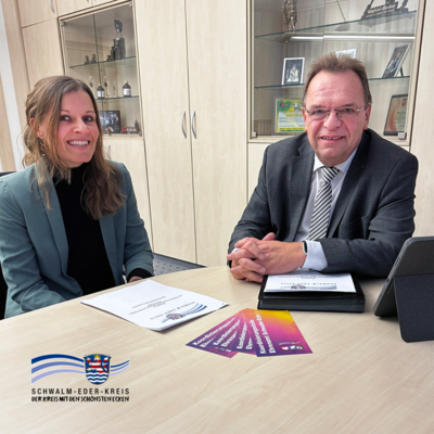 In einem Büro sitzen eine Frau und ein Mann an einem Besprechungstisch und lächeln in die Kamera. Auf dem Tisch liegen Unterlagen mit dem Logo des Schwalm-Eder-Kreises sowie mehrere farbige Flyer; rechts steht ein Tablet in einer Hülle. Im Hintergrund sind helle Schränke und eine Vitrine zu sehen, unten links ist das Kreis-Logo eingeblendet.