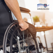 Nahaufnahme einer Person im Rollstuhl. Eine Hand greift an das große Rad des Rollstuhls, um sich fortzubewegen. Im Hintergrund ist ein heller Innenraum mit Tisch und unscharf erkennbaren Möbeln zu sehen. Oben rechts befindet sich das Logo des Schwalm-Eder-Kreises.