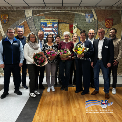 Gruppenbild mit mehreren Mitarbeitenden und Geehrten vor einer großen Wappenwand des Schwalm-Eder-Kreises. Einige Personen halten Blumensträuße in den Händen. Das Signet des Schwalm-Eder-Kreises ist im Bild eingeblendet.