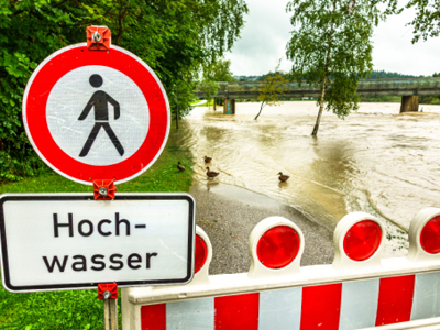 as Bild zeigt eine überflutete Uferzone, bei der der Weg entlang eines Flusses vollständig unter Wasser steht. Im Vordergrund befindet sich eine rot-weiße Absperrung mit einem Verbotsschild für Fußgänger und einem zusätzlichen Schild mit der Aufschrift „Hochwasser“. Im Hintergrund sind Bäume, eine Brücke und mehrere Enten im Wasser zu sehen. Die Szene verdeutlicht die Gefahr durch Starkregen und Überschwemmung.