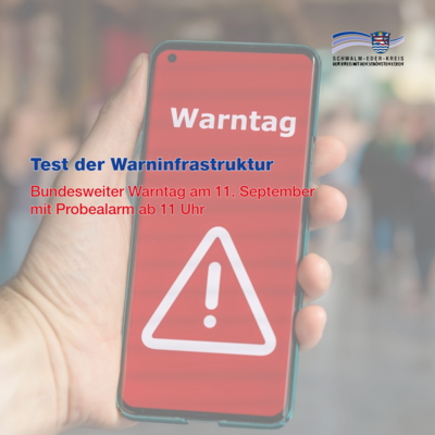 Eine Hand hält ein Smartphone mit rotem Bildschirm, auf dem in weiß ein Warndreieck mit Ausrufezeichen und der Schriftzug „Warntag“ zu sehen ist. Darüber liegt Text: „Test der Warninfrastruktur – Bundesweiter Warntag am 11. September mit Probealarm ab 11 Uhr“. Oben rechts das Logo des Schwalm-Eder-Kreises.