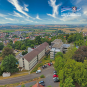 Auf dem Bild ist die Kreisverwaltung des Schwalm-Eder-Kreises in der Parkstraße in Homberg (Efze) aus der Luftperspektive zu sehen. Die Verwaltungsgebäude liegen zwischen vielen Bäumen, Parkplätzen und umliegenden Wohnhäusern. Im Hintergrund erstreckt sich eine weite Landschaft mit Feldern, Dörfern und einem klaren blauen Himmel mit hohen, weißen Wolken. Oben rechts befindet sich das Logo des Schwalm-Eder-Kreises.