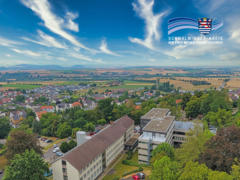 Auf dem Bild ist die Kreisverwaltung des Schwalm-Eder-Kreises in der Parkstraße in Homberg (Efze) aus der Luftperspektive zu sehen. Die Verwaltungsgebäude liegen zwischen vielen Bäumen, Parkplätzen und umliegenden Wohnhäusern. Im Hintergrund erstreckt sich eine weite Landschaft mit Feldern, Dörfern und einem klaren blauen Himmel mit hohen, weißen Wolken. Oben rechts befindet sich das Logo des Schwalm-Eder-Kreises.