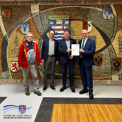 Vor der großen Mosaikwand mit den Stadt- und Gemeindewappen des Schwalm-Eder-Kreises stehen vier Männer nebeneinander. Von links sind zu sehen: Volker Baranowski vom Kreisstraßenbau des Schwalm-Eder-Kreises, Christoph Sippel (Mitglied des Hessischen Landtags, Bündnis 90/Die Grünen), Knut John (Landesbeauftragter für ländliche Räume) und Jürgen Kaufmann (Vize-Landrat des Schwalm-Eder-Kreises). Knut John und Jürgen Kaufmann halten gemeinsam einen Förderbescheid in den Händen. Links unten ist das Logo des Schwalm-Eder-Kreises eingeblendet.