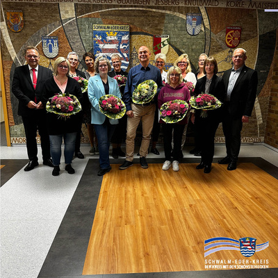 Gruppenfoto im Landratsamt des Schwalm-Eder-Kreises. Vor einem Mosaik mit Kreiswappen stehen dreizehn Personen in zwei Reihen. In der vorderen Reihe halten acht Personen Blumensträuße in den Händen – sie wurden geehrt oder verabschiedet. Ganz links im Bild steht Landrat Winfried Becker, ganz rechts Erster Kreisbeigeordneter Jürgen Kaufmann. Die weiteren abgebildeten Personen sind (von links nach rechts): Katrin Ellenberger, Sandra Mausehund, Gabi Deiseroth, Kathrin Wissemann, Christiane Vollbrecht, Jan Siebert, Manuela Zülch, Petra Ziepprecht, Claudia Asbrand, Angelika Roth und Julia Mertens. Unten rechts im Bild ist das Logo des Schwalm-Eder-Kreises zu sehen mit dem Schriftzug: „Der Kreis mit den schönsten Ecken“.