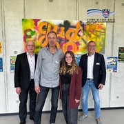Vier Personen stehen vor einer Betonwand.  Auf der Wand hängt ein buntes Graffiti-Kunstwerk mit großen orangefarbenen Buchstaben. Unten rechts ist das Logo „Schwalm-Eder-Kreis – Der Kreis mit den schönsten Ecken“ zu sehen.