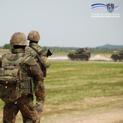 Das Bild zeigt zwei Bundeswehrsoldaten in voller Ausrüstung, die auf einem Truppenübungsplatz unterwegs sind. Im Hintergrund sind zwei gepanzerte Fahrzeuge in Bewegung zu sehen. Die Szene spielt sich auf einer offenen Grasfläche mit leicht hügeliger Landschaft ab. Auch hier ist das Logo des Schwalm-Eder-Kreises mit dem Schriftzug „Der Kreis mit den schönsten Ecken“ im Bild oben rechts eingefügt.