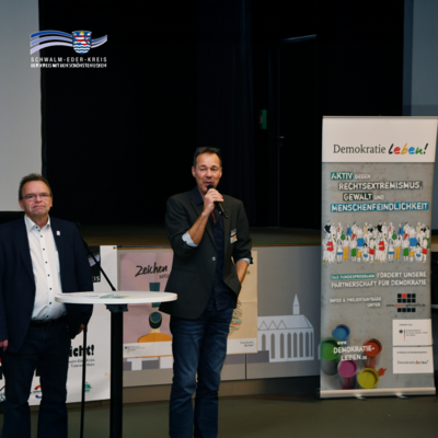 Auf dem Bild stehen Landrat Winfried Becker (links) und Tom Werner (Projektkoordinator Gewalt geht nicht!) in einem Saal auf einer Bühne. Werner spricht in ein Mikrofon, Becker steht daneben an einem runden Stehtisch. Im Hintergrund ist ein Banner mit der Aufschrift „Demokratie leben!“ zu sehen, das auf das Bundesprogramm gegen Rechtsextremismus, Gewalt und Menschenfeindlichkeit hinweist. Links im Bild sind weitere Plakate mit dem Schriftzug „Zeichen setzen!“ erkennbar. Oben links befindet sich das Logo des Schwalm-Eder-Kreises mit dem Slogan „Der Kreis mit den schönsten Ecken“.